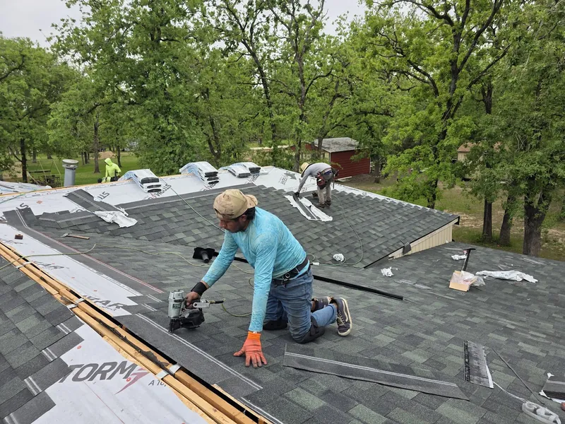 Roofer kneeling on a roof nailing down new gray architectural shingles over synthetic underlayment