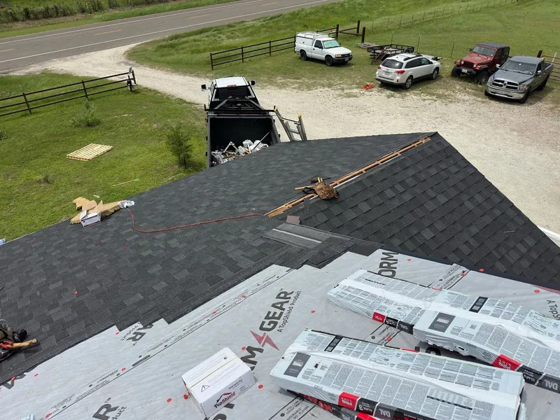 New dark shingles going on over synthetic underlayment, with underlayment rolls staged on the roof and work trucks in the drive